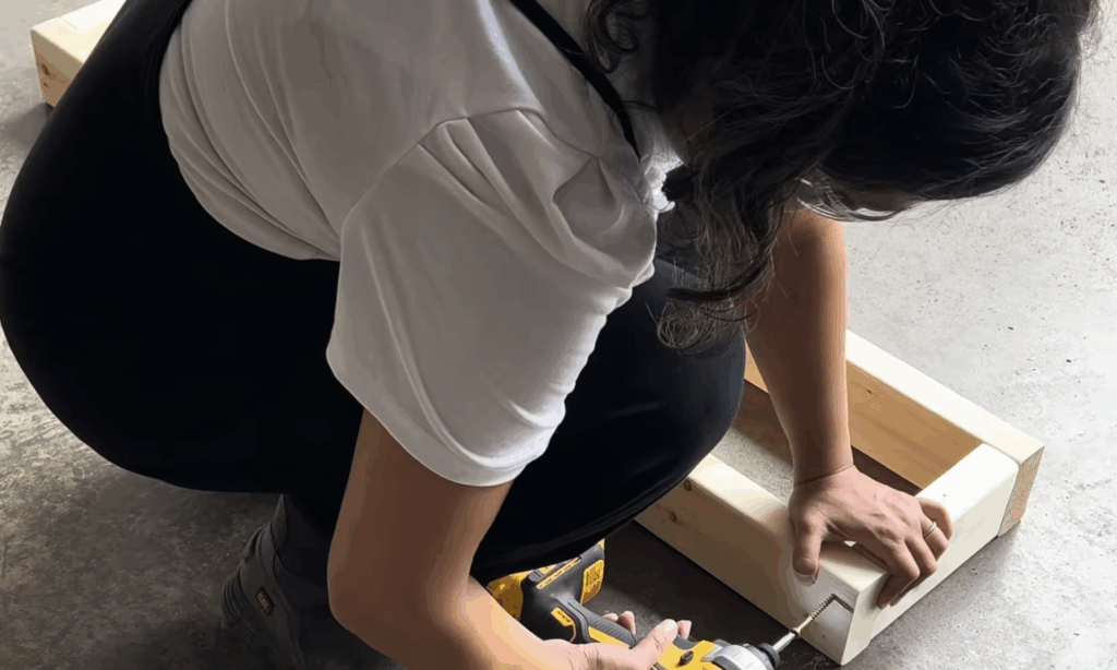 Assembling the wooden fireplace frame on the garage floor using a Dewalt drill and 2x4 boards.