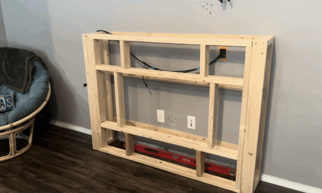 Finished wooden frame for a small electric fireplace installed against a grey wall with power cables routed inside.