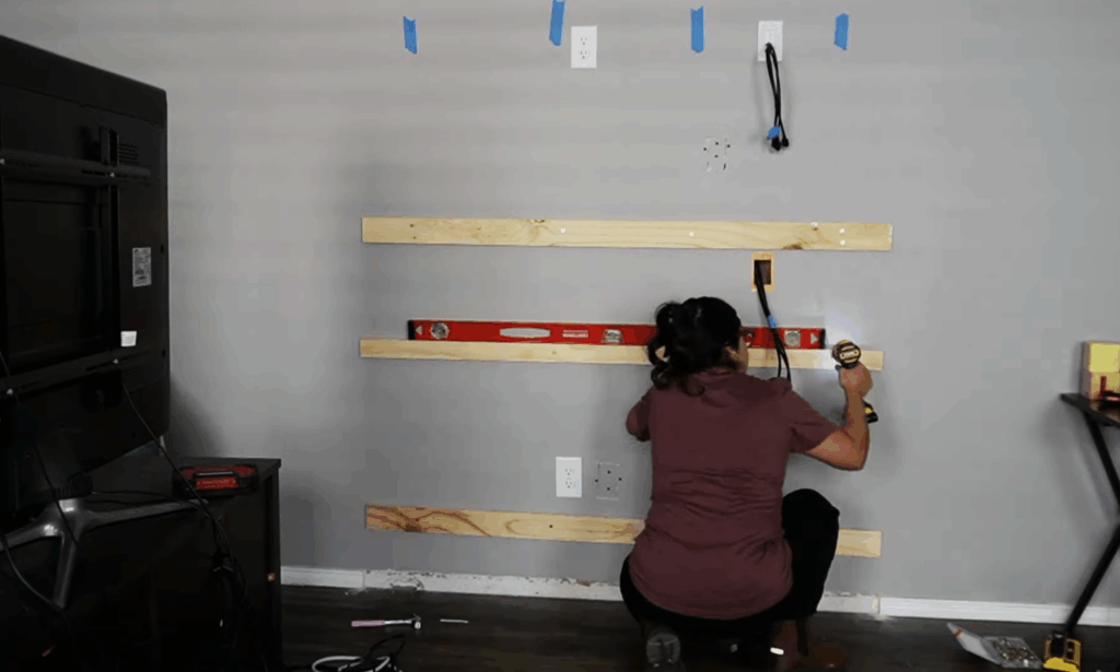 Installing horizontal 2x4 boards on a grey wall to anchor the new fireplace frame, using a drill and level for alignment.