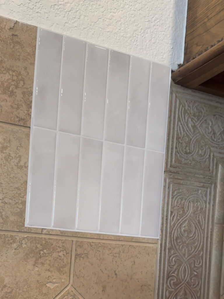 Side-by-side view showing old beige tile next to new white peel-and-stick tile during kitchen backsplash update.