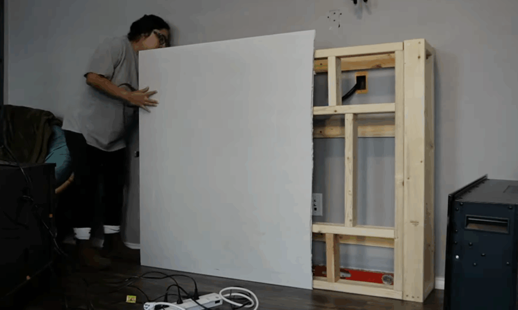 Positioning a large drywall sheet to cover the DIY fireplace structure, preparing for final wall coverage.