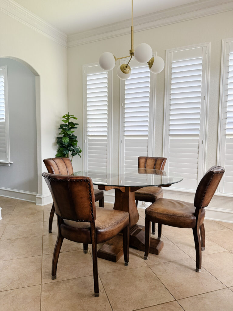 Bright dining nook with round glass table, leather chairs, new modern brass light fixture, and white plantation shutters
