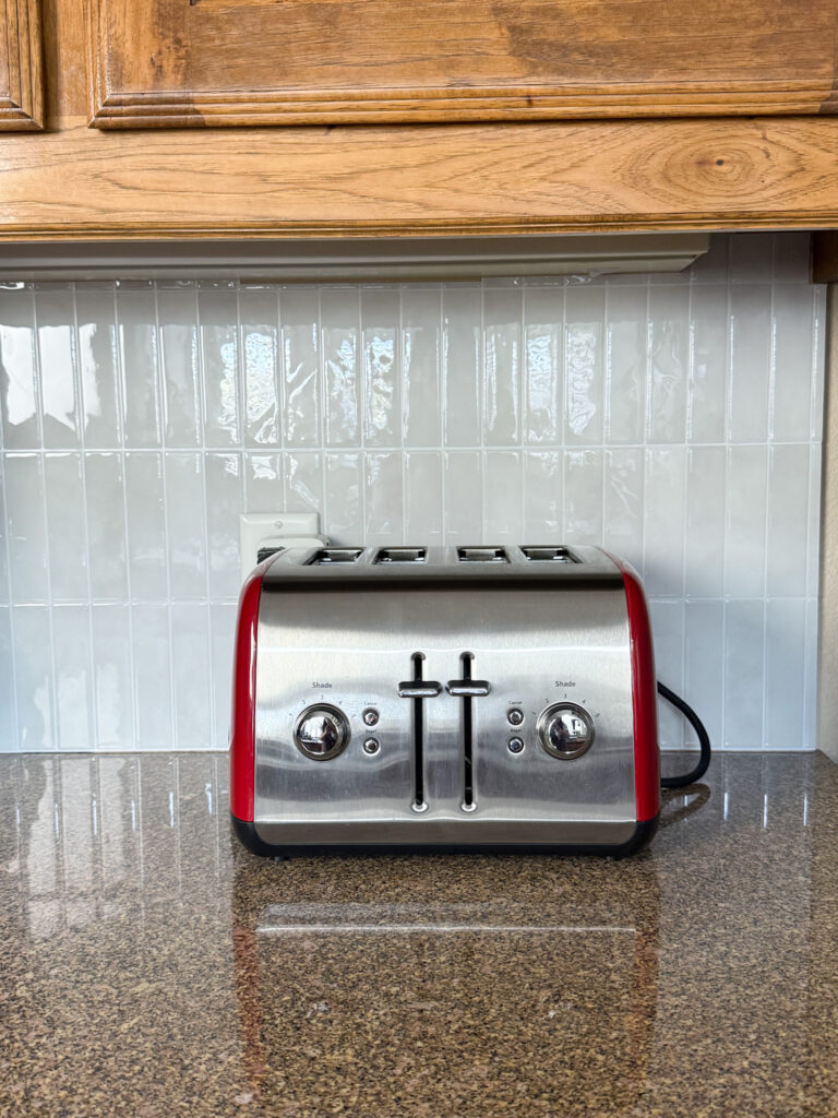 Section of updated kitchen with wooden cabinets and glossy peel-and-stick tile backsplash behind a red toaster.