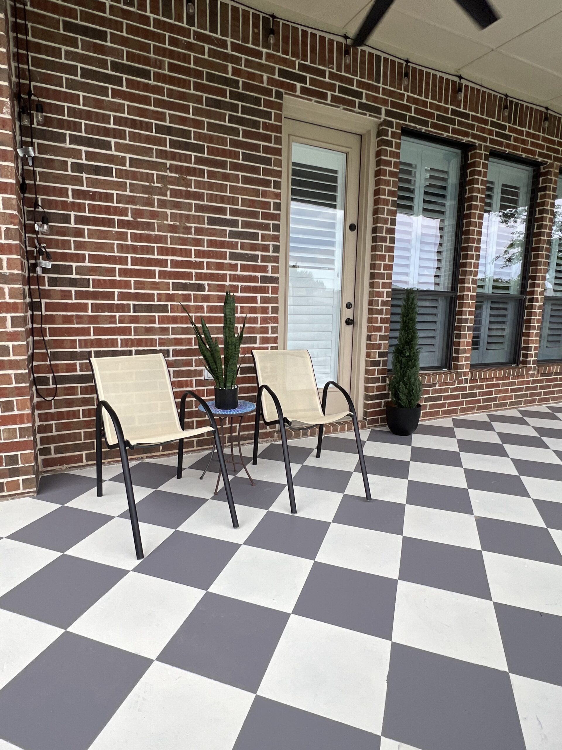 Checkered patio with two light chairs and a small table against the brick wall.