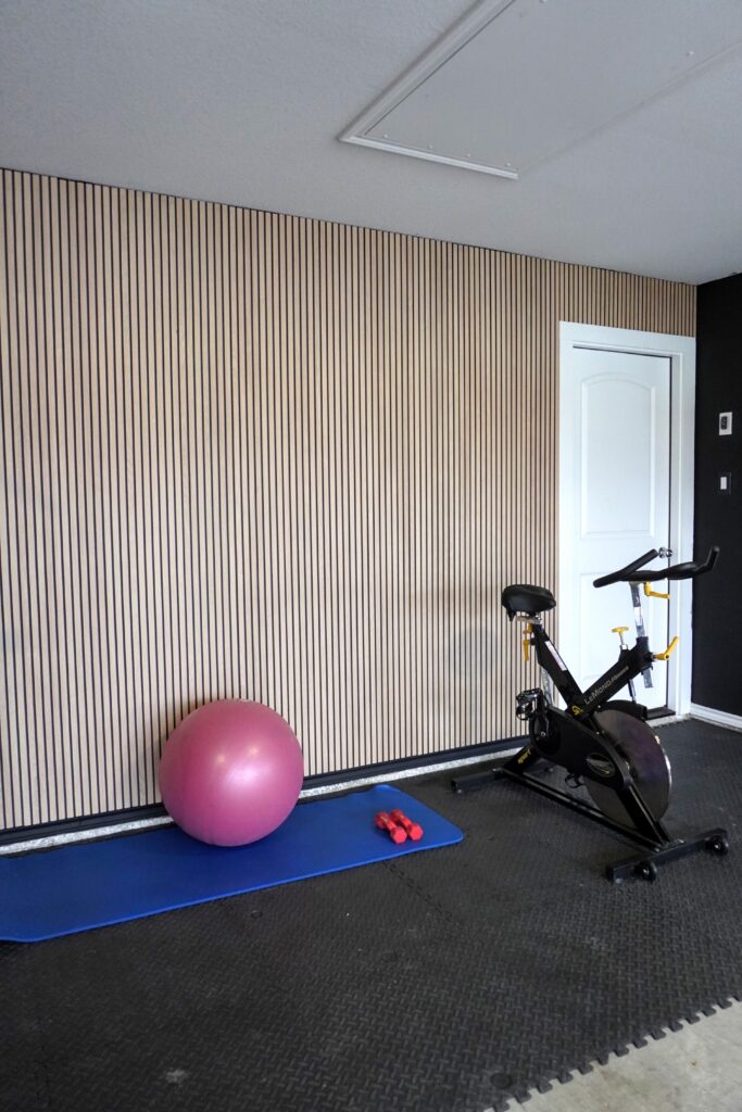 DIY fluted wall panel accent wall in a home gym showing full height installation with warm oak MDF panels, black rubber flooring, and white door
