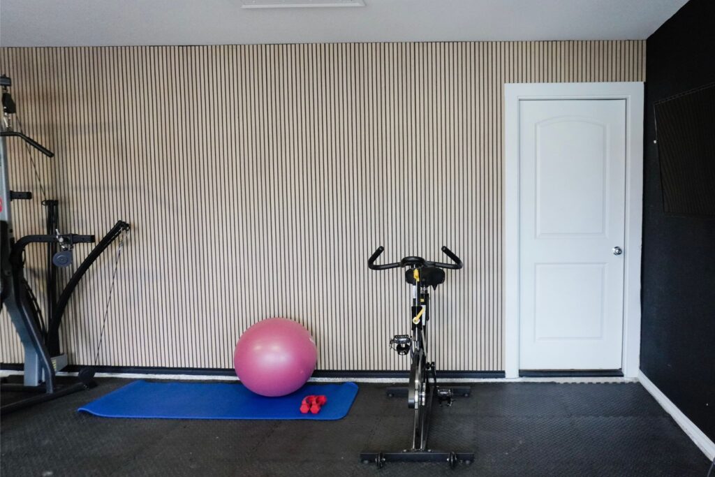 Full view of a DIY fluted MDF panel accent wall in a home gym with warm oak finish and black slats, spin bike and yoga mat in front