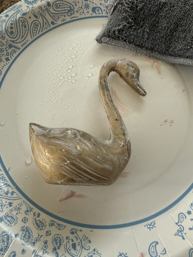 Brass swan being cleaned with The Pink Stuff paste in a bowl with a microfiber cloth showing the cleaning process