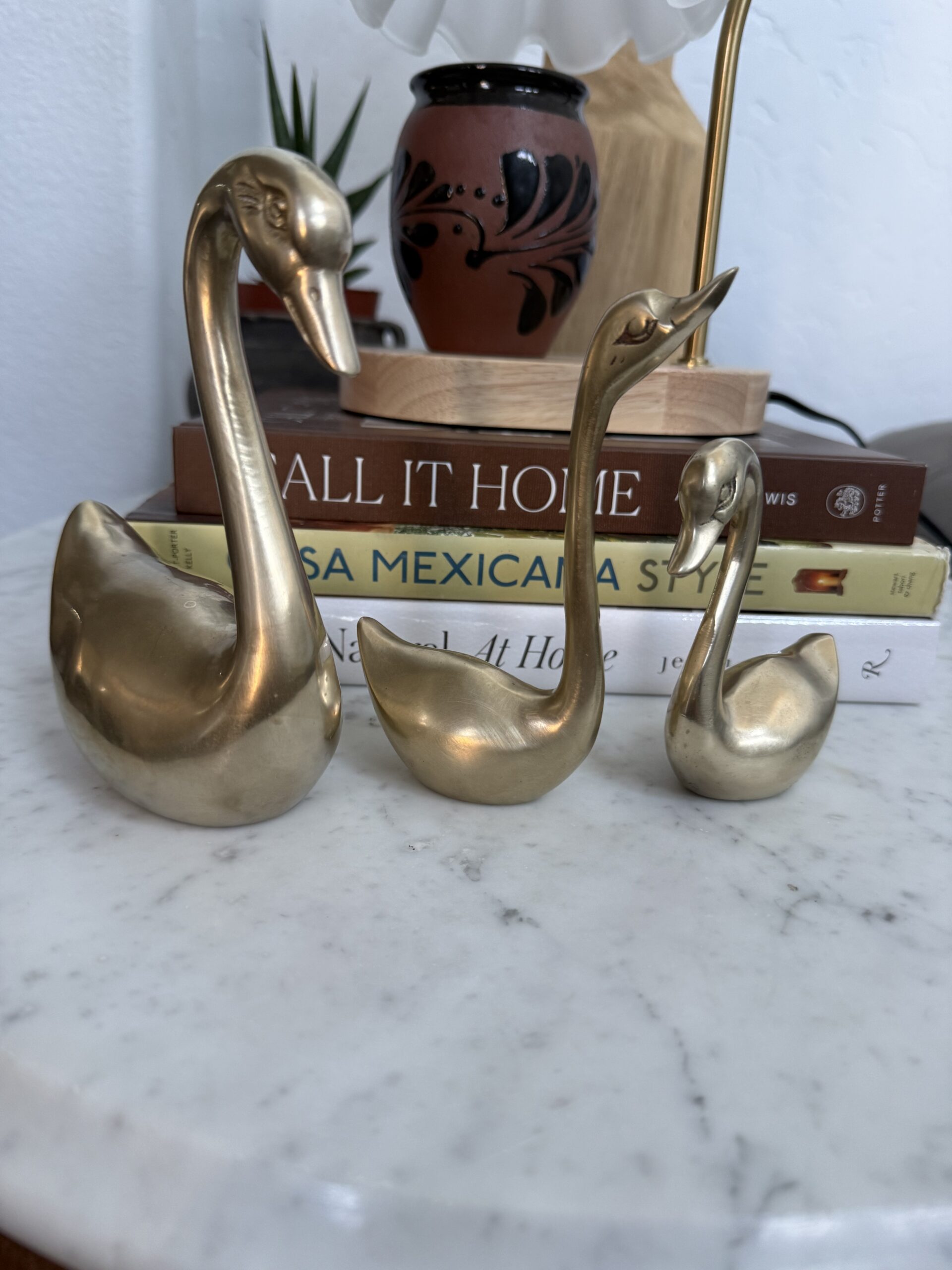 Three polished brass swans of different sizes displayed on a marble surface with Casa Mexicana Style and other books