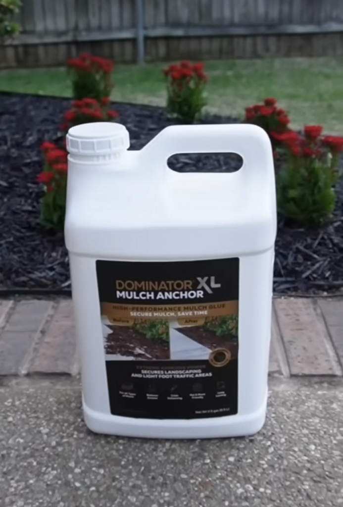 an XL bottle of Dominator Mulch Anchor sitting next to a flower bed in a yard.